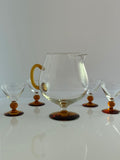 A clear glass carafe with a rounded body, a curved amber handle, and a dark amber foot, shown with five matching small goblets on a glossy white surface against a light gray background.