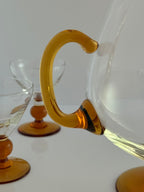 A close-up of a clear glass carafe with a thick, curved amber-yellow handle, shown alongside matching clear and amber glassware on a white surface against a light gray background.
