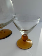 A clear stemmed cocktail coupe glass with a round amber-colored base, shown on a white surface with another similar glass partially visible in the background.