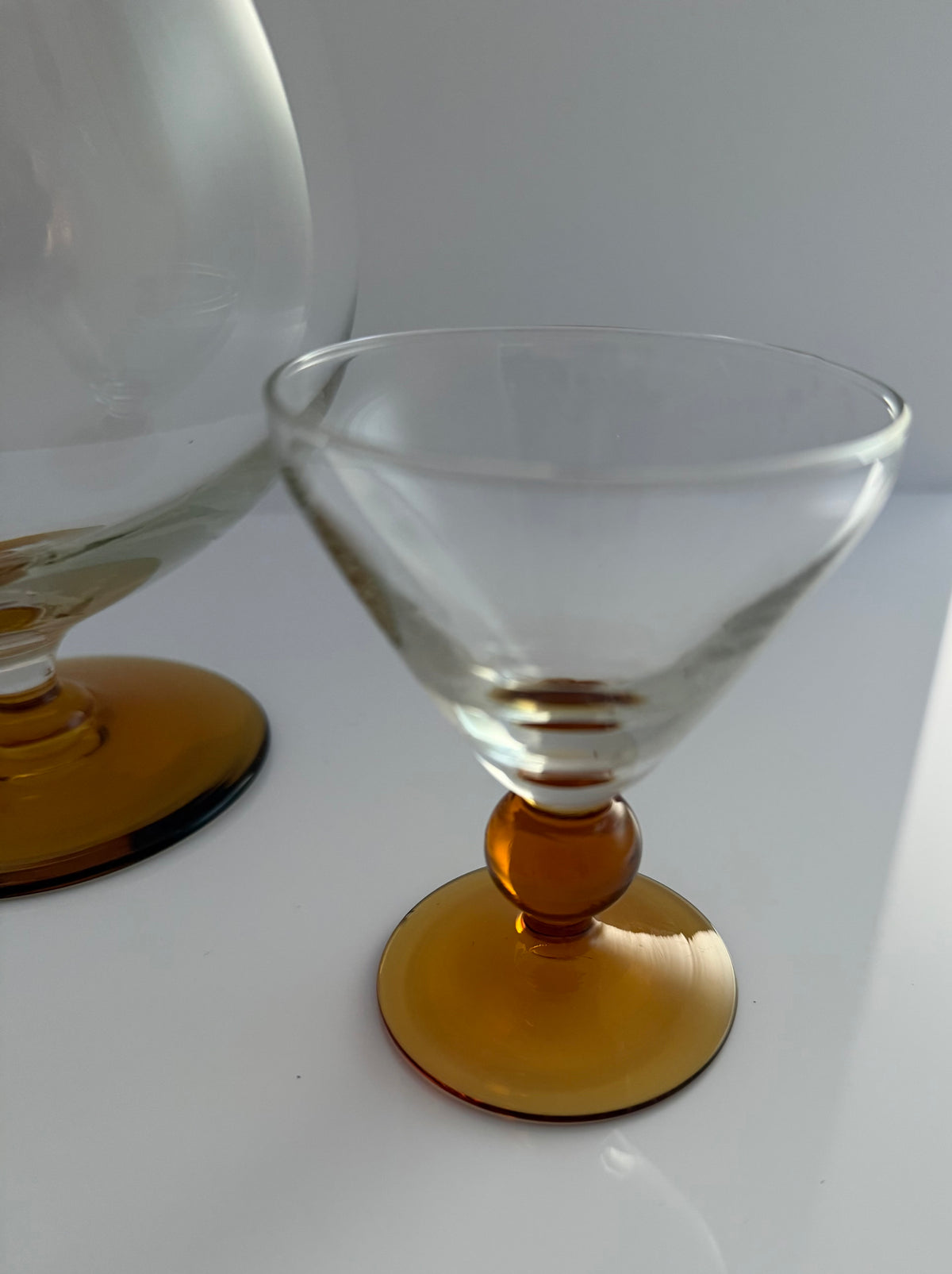 A clear stemmed cocktail coupe glass with a round amber-colored base, shown on a white surface with another similar glass partially visible in the background.