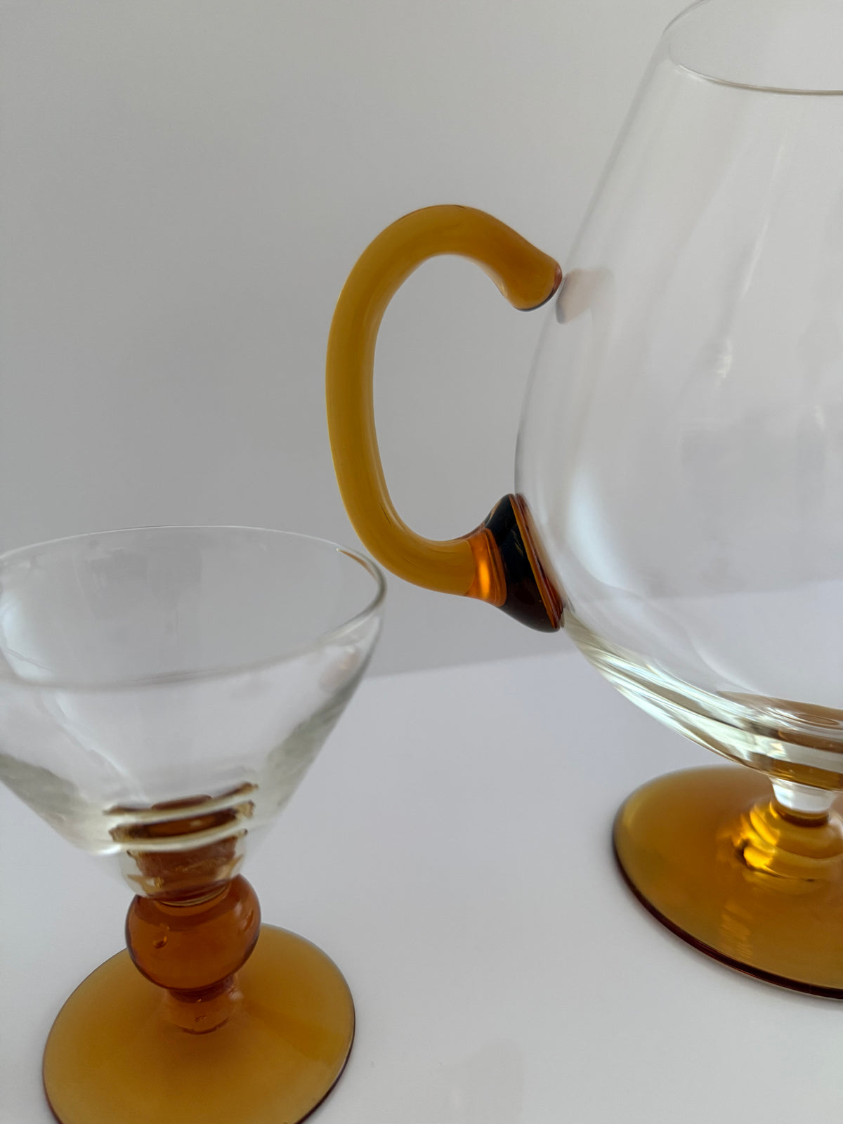 A clear glass carafe with a curved amber-yellow handle and matching circular base, shown next to a matching clear glass coupe-style tumbler on a white surface against a light gray background.