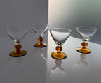 Four clear glass cocktail coupe glasses with amber-colored bases, arranged on a reflective white surface against a gray background.