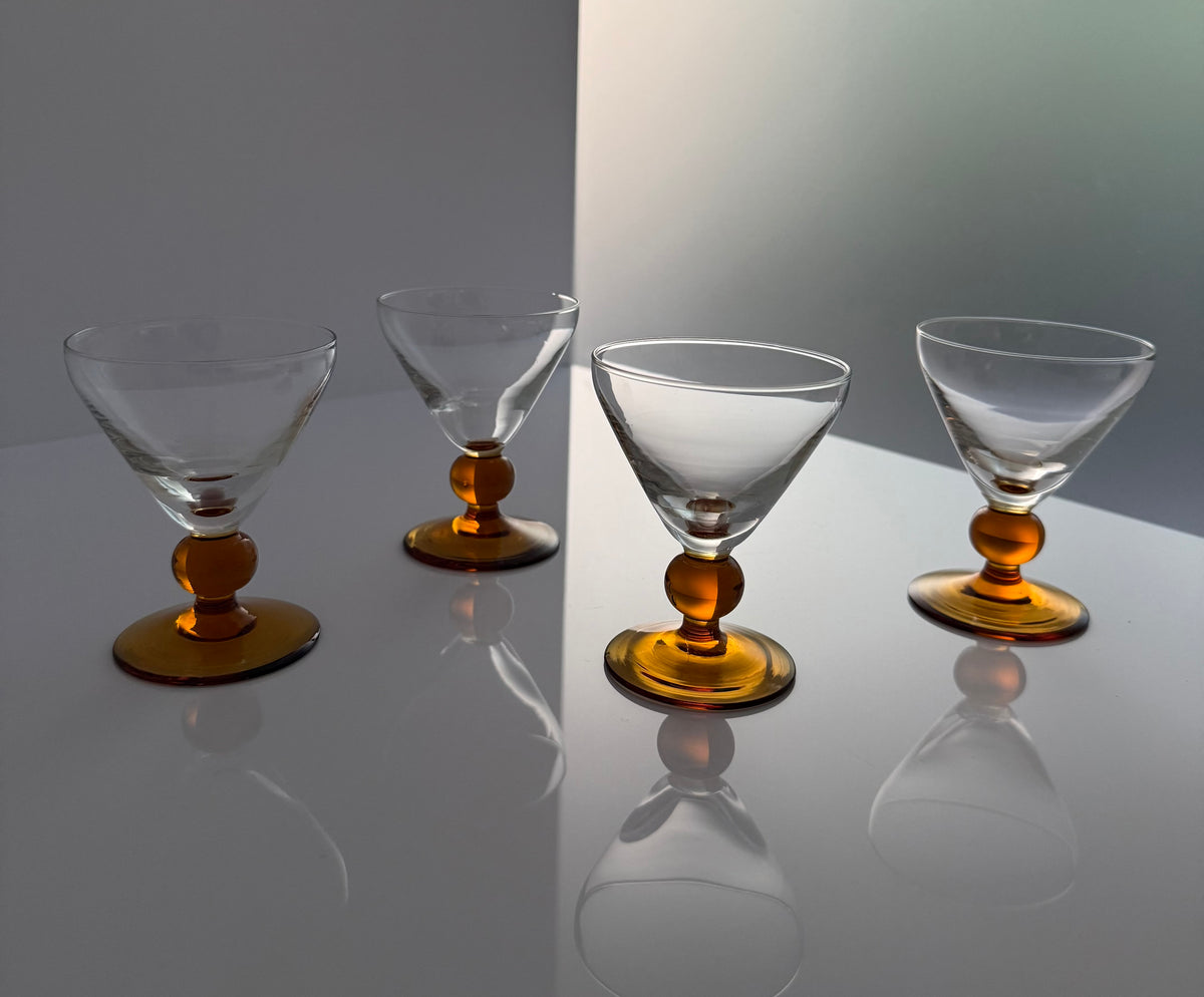 Four clear glass cocktail coupe glasses with amber-colored bases, arranged on a reflective white surface against a gray background.