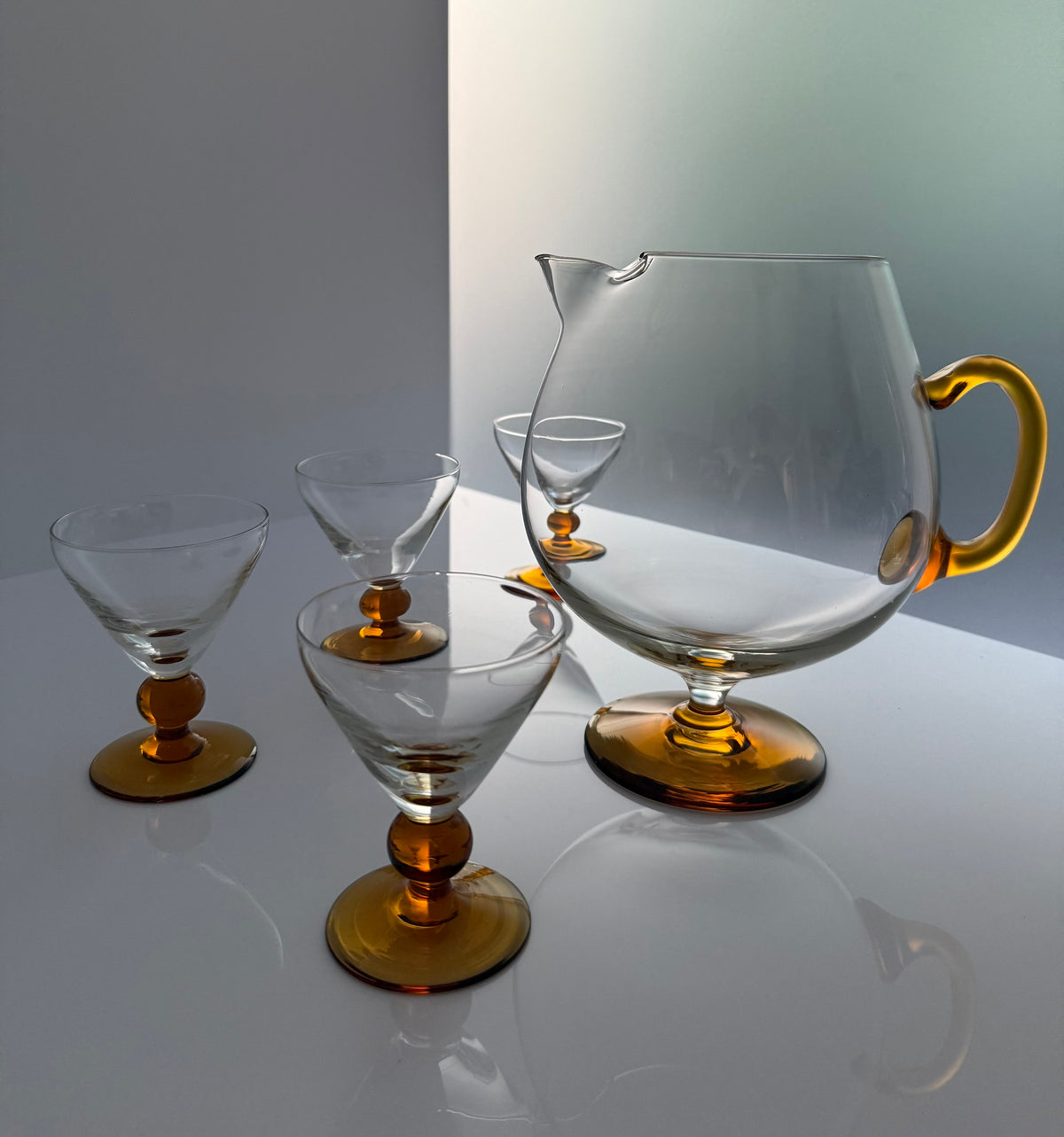 A set of clear glass drinking vessels with amber-colored bases and handles, including a large pitcher and four stemmed glasses, arranged on a reflective white surface.