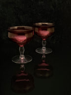 Two vintage-style coupe glasses with faceted dark red glass bowls and clear stems, set against a dark reflective background.