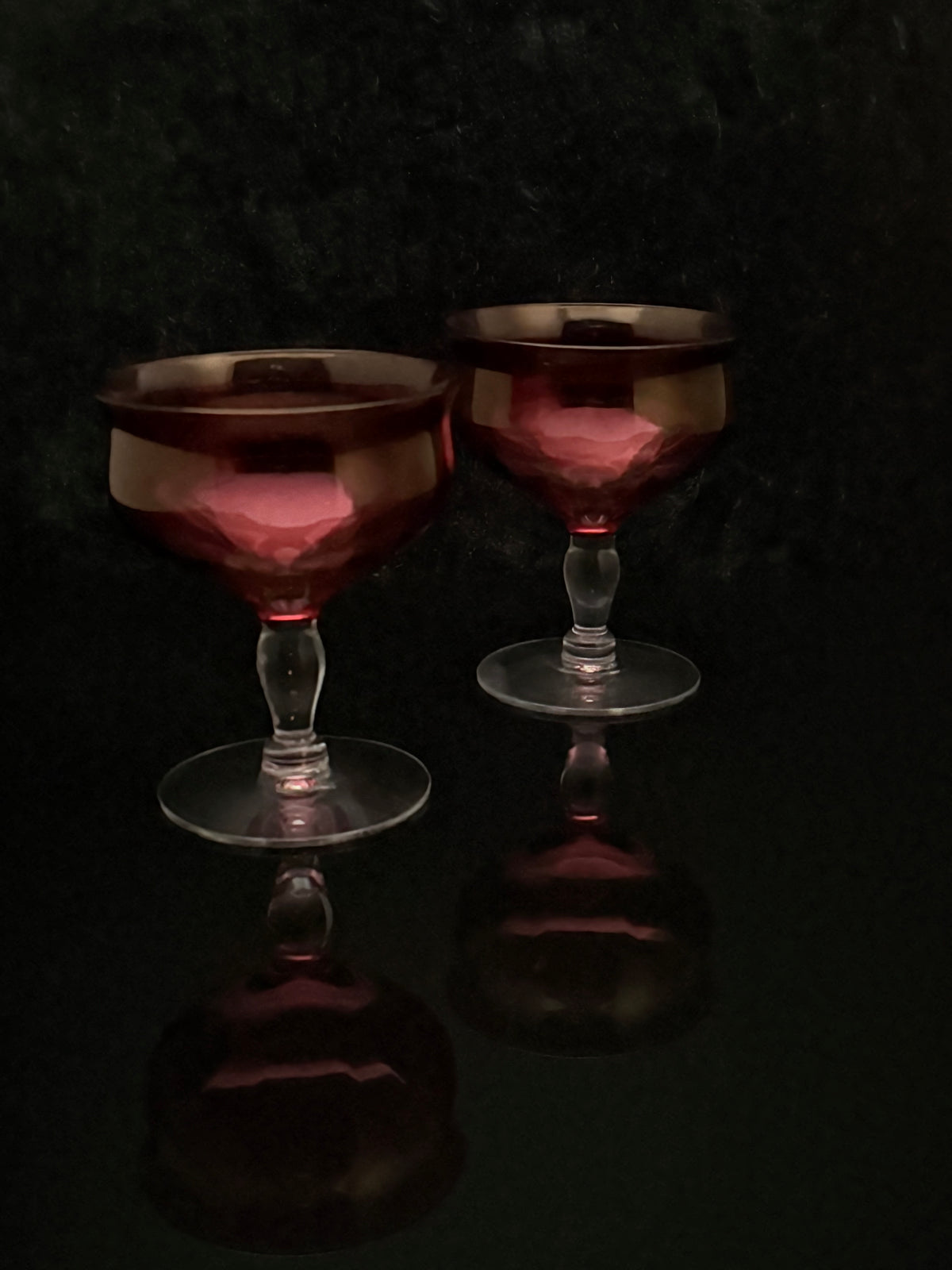 Two vintage-style coupe glasses with faceted dark red glass bowls and clear stems, set against a dark reflective background.