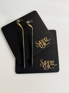 Two steel cocktail picks with gold-toned accents, displayed on black genuine leather branded coasters with gold 'Sipply Chic' lettering, on a white background.