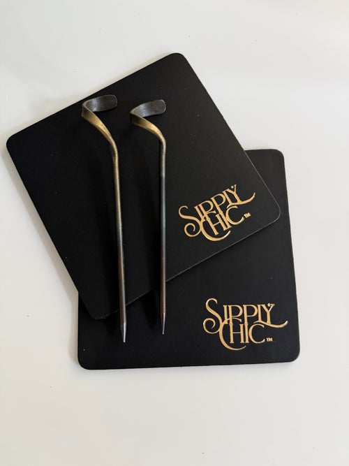 Two steel cocktail picks with gold-toned accents, displayed on black genuine leather branded coasters with gold 'Sipply Chic' lettering, on a white background.