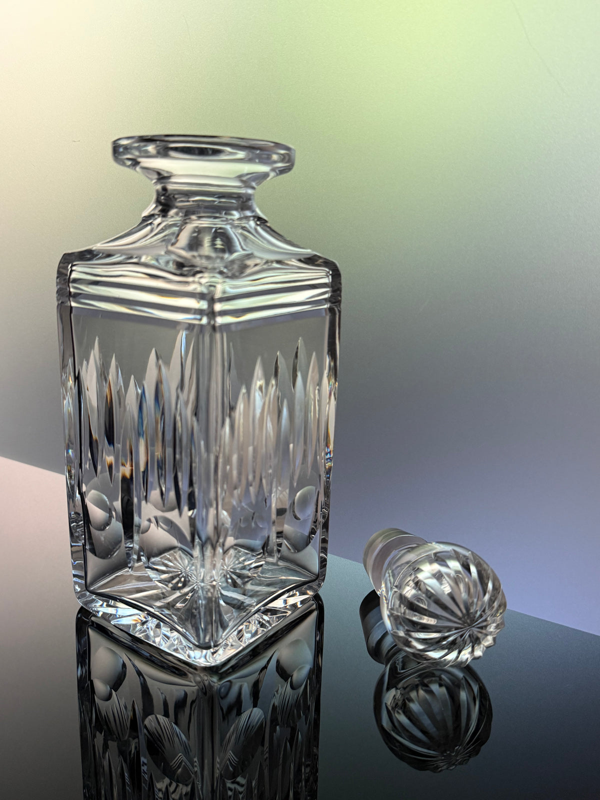 A clear cut-glass decanter with a matching stopper, shown on a reflective dark surface against a softly lit gradient background.