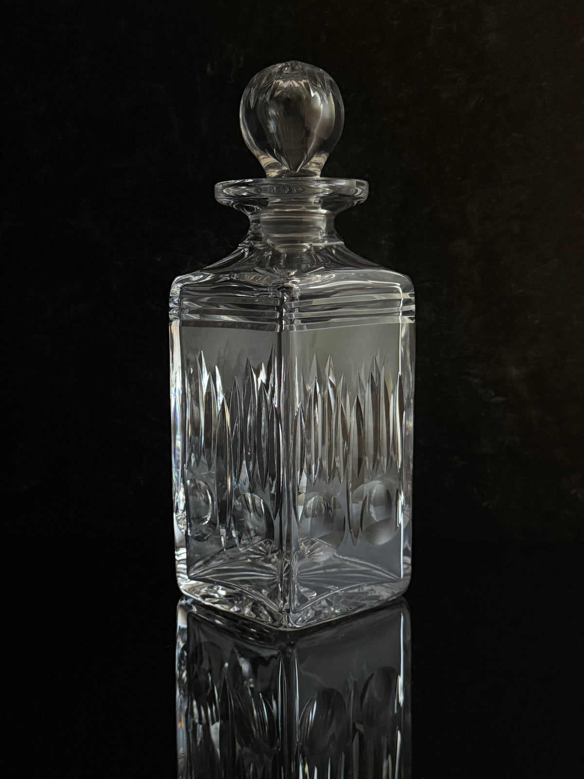 A clear cut-glass decanter with a matching round stopper, photographed against a dark reflective background.