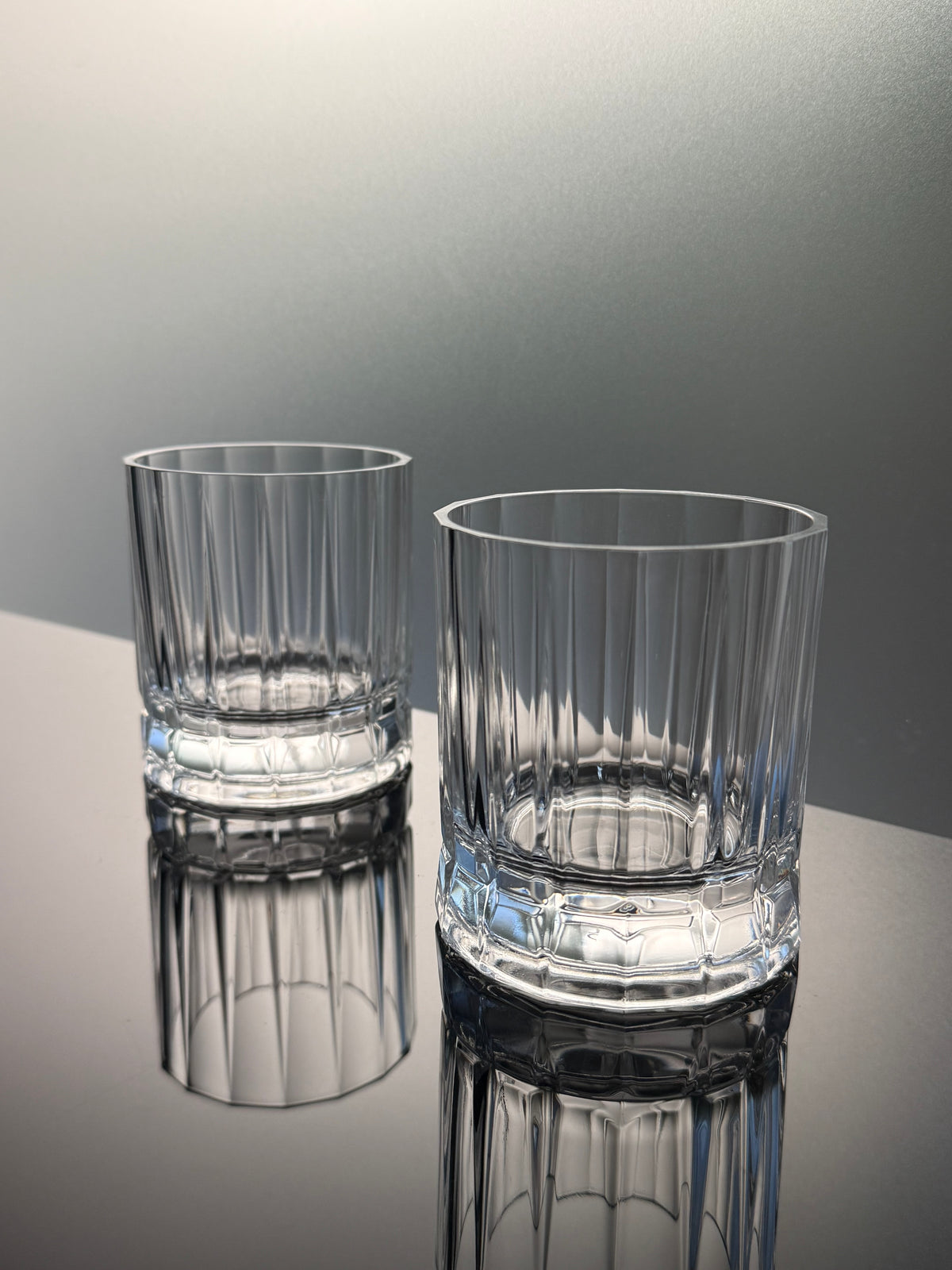 Two clear cut-glass rocks tumblers with vertical fluting, displayed on a reflective dark surface against a gray gradient background.
