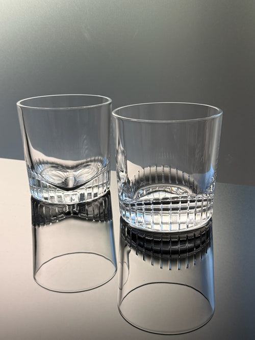 Two clear cut-glass rocks tumblers with a textured, ribbed base, shown on a reflective surface against a gray background.