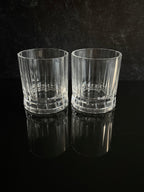 Two clear ribbed whiskey glasses with faceted bases, standing side by side on a glossy black reflective surface against a dark textured background.