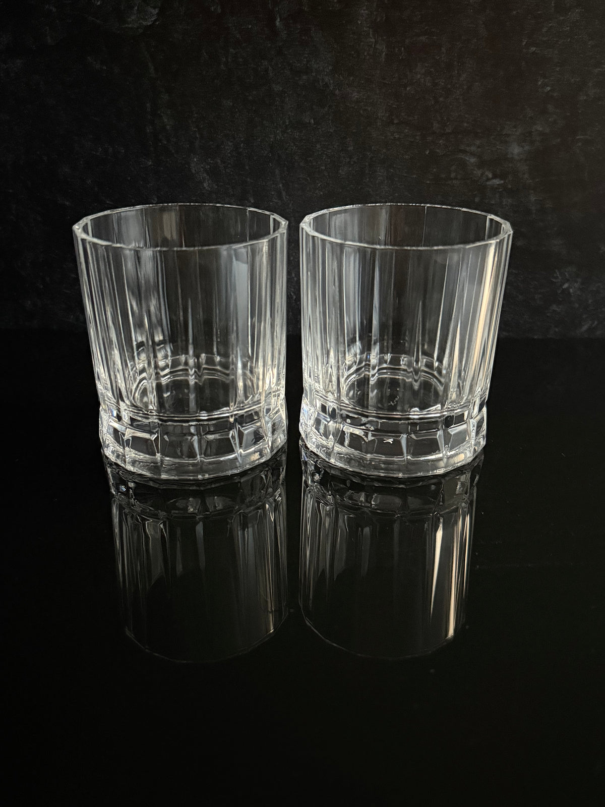 Two clear ribbed whiskey glasses with faceted bases, standing side by side on a glossy black reflective surface against a dark textured background.