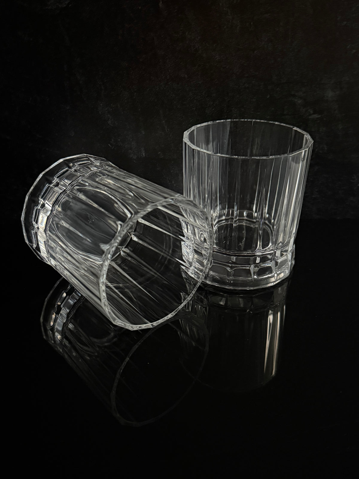 Two clear ribbed glass tumblers on a glossy black reflective surface against a dark background, with one glass standing upright and the other lying on its side.