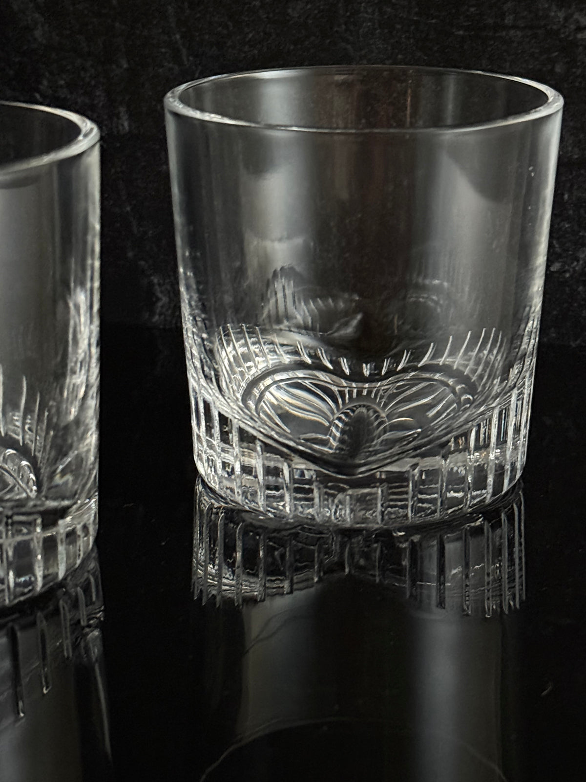 A close-up of a clear cut-glass tumbler with a textured patterned band, shown against a dark background.